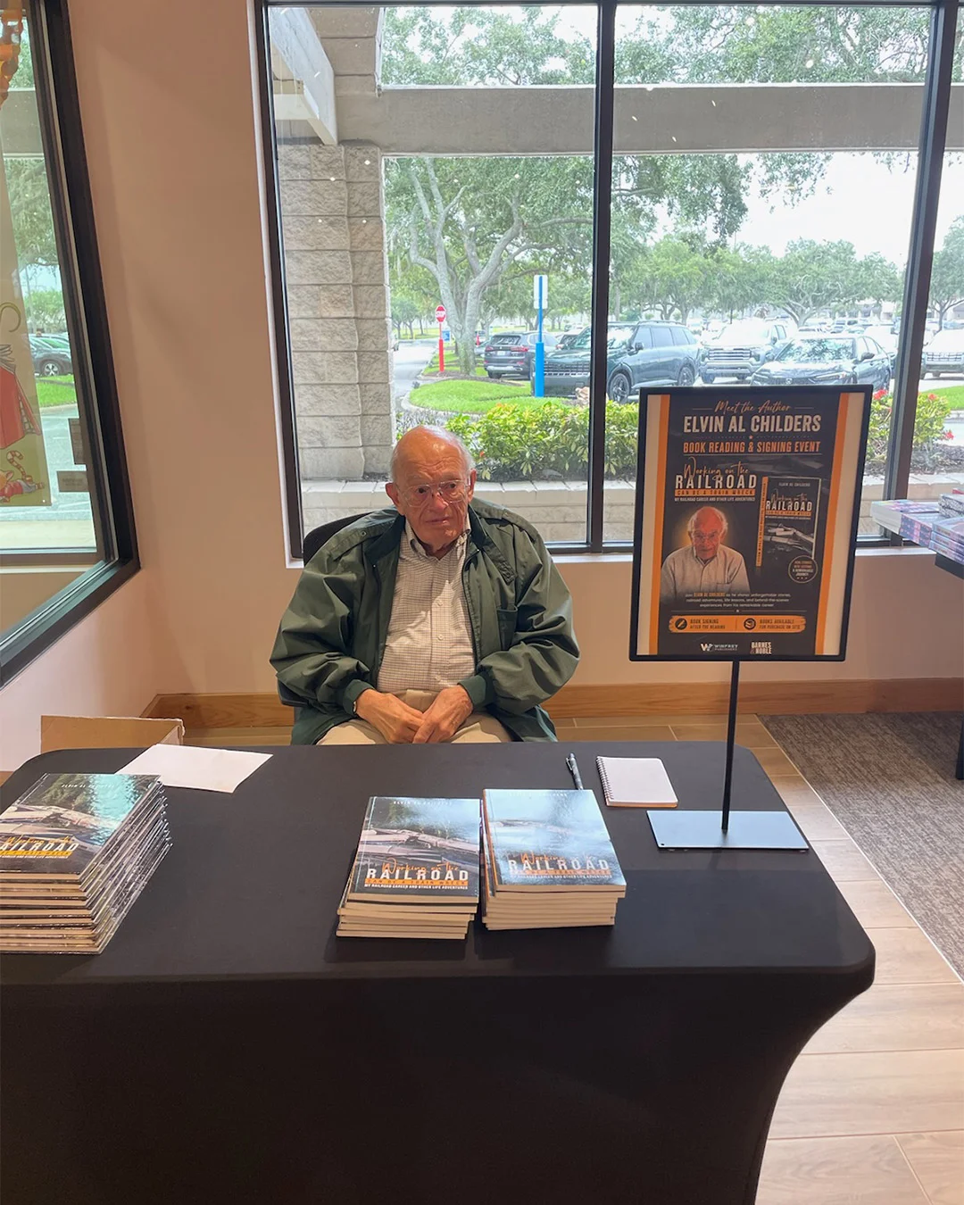 An author seated at a signing table by the shop window, stacked copies of his book in front of him and a meet-the-author poster on an easel alongside