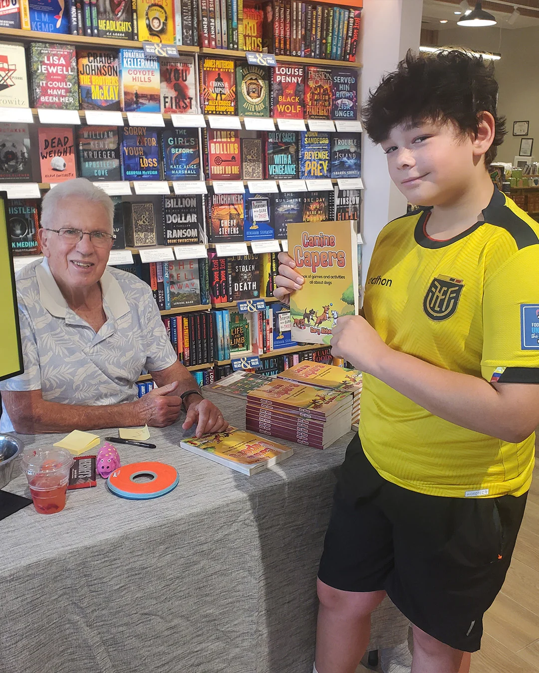 An author at his signing table beside a young reader holding up a copy of the book, in front of a wall of new releases