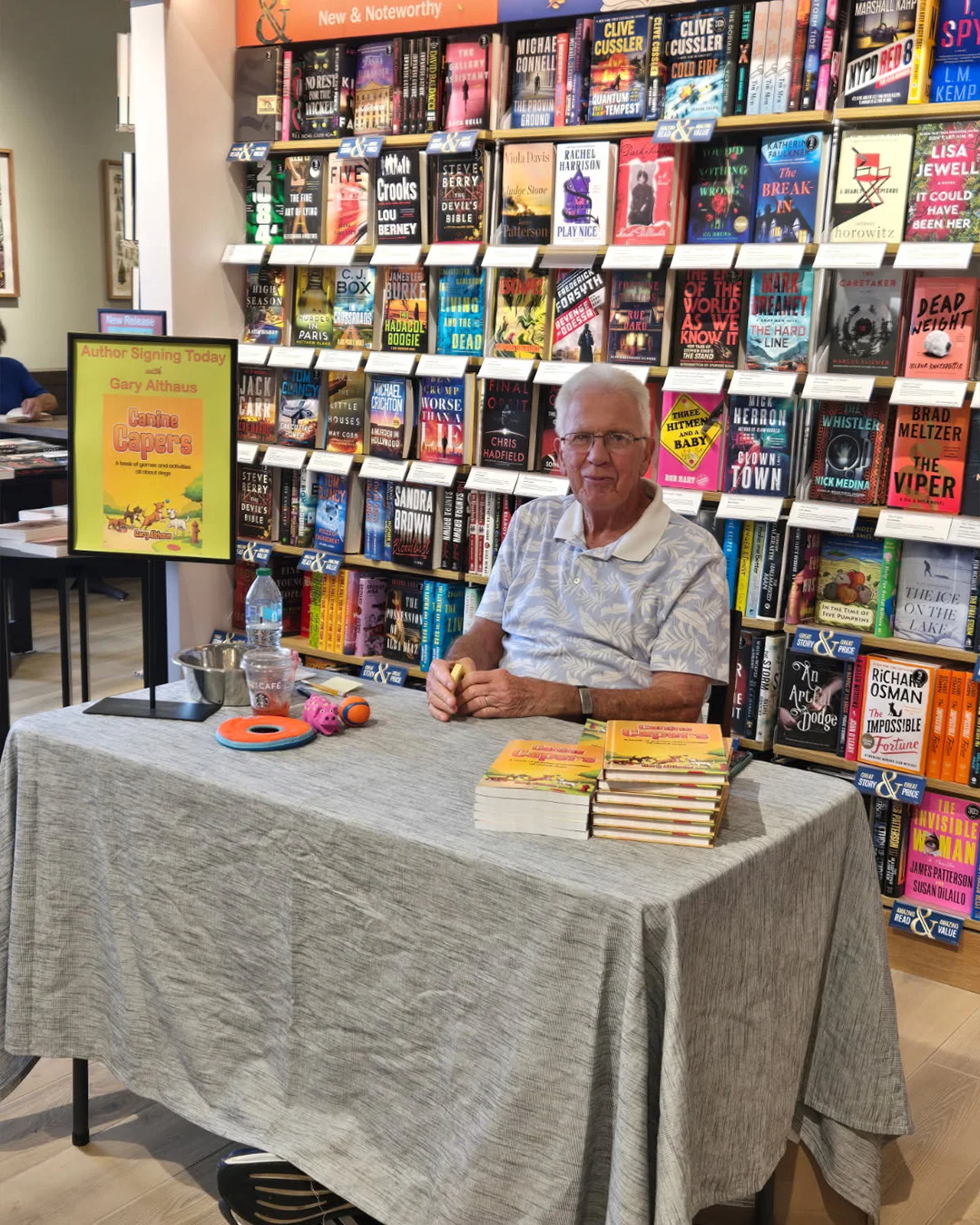 An author waiting at a draped signing table, an author-signing-today poster on an easel and copies stacked ready in front of him