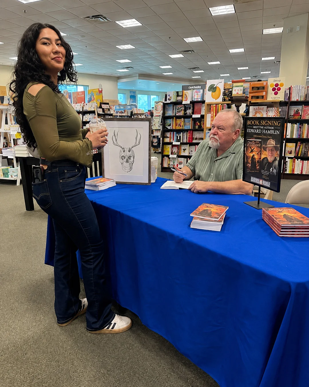 A reader waiting beside the signing table while the author signs, the framed cover artwork standing on the table